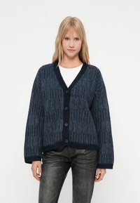 O femeie tânără îmbrăcată cu un cardigan în dungi de culoare albastru închis, cu butoni, o bluză albă și pantaloni negri texturați, stând în fața unui fundal simplu.