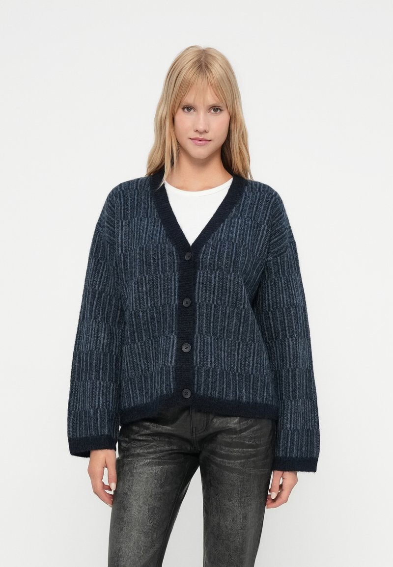 O femeie tânără îmbrăcată cu un cardigan în dungi de culoare albastru închis, cu butoni, o bluză albă și pantaloni negri texturați, stând în fața unui fundal simplu.