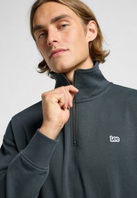 Fleece zip-up grigio scuro con colletto alto, caratterizzato da un piccolo logo bianco "Lee" sul petto e dettagli in tessuto testurizzato.