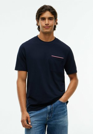 Junger Mann mit mittellangen dunklen Haaren, der ein dunkelblaues T-Shirt mit einer kleinen Brusttasche und blaue Jeans trägt, steht mit einer Hand in der Tasche.
