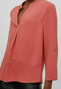 Blusa corallo in tessuto testurizzato, con scollatura a V e maniche a tre quarti. Presenta un dettaglio a bottone sulla scollatura e una vestibilità comoda.