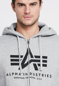 Šedá mikina na zips vyrobená z zmesi bavlny, s čiernym logom so štýlizovaným písmenom "A" a pruhmi. Kapucňa s nastaviteľným šnúrkou.