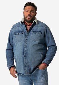Camicia di jeans con un lavaggio azzurro chiaro, due tasche sul petto e un morbido colletto bianco. Indossata sopra una camicia a quadri rossa, mostrata in posizione eretta.