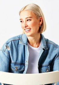 Giubbotto di denim con dettagli invecchiati, colore azzurro chiaro, chiusura a bottoni, indossato sopra una maglietta bianca a coste. Capelli biondi corti, sfondo chiaro.