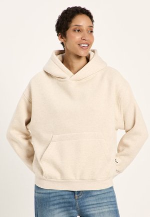 Person mit kurzen lockigen Haaren, die einen beigen Kapuzenpullover und blaue Jeans trägt, lächelt und sieht zur Seite, die Hände hinter dem Rücken.
