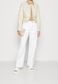 Veste en cuir beige courte avec fermeture à boutons, haut beige, pantalon blanc à jambes larges et sandales à plateforme blanches. Comprend des détails métalliques minimalistes.