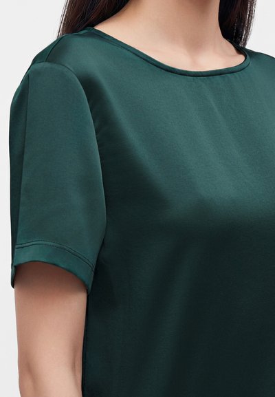Femme portant un chemisier vert foncé à manches courtes avec un col rond, laissant apparaître les épaules et le haut de la poitrine.