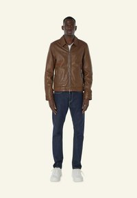 Bruine leren jas met een kraag, voorkleurige ritssluiting en zijzakken, gecombineerd met donkere denim jeans en witte sneakers. Effen achtergrond.