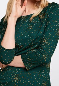 Mörkgrön klänning med trekvartslånga ärmar, prydd med ett spritt mönster av guldprickar och en rundad halsringning. Tillverkad av ett mjukt, texturerat tyg.