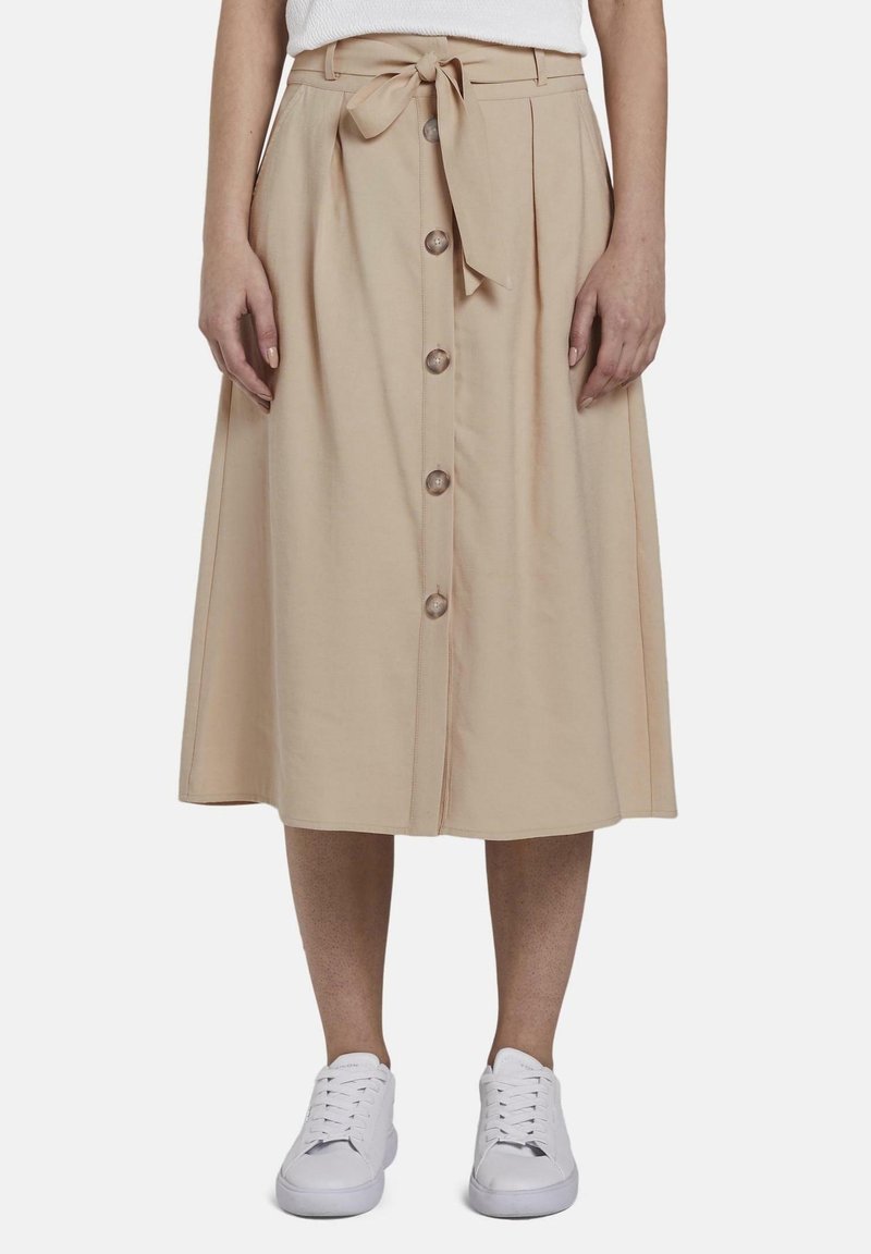 Jupe midi beige avec boutons sur le devant et ceinture à la taille, portée avec des baskets blanches et un top blanc rentré, montrée de la taille aux pieds.