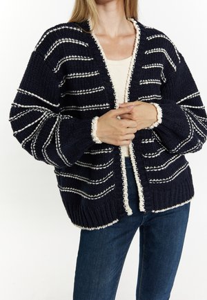 Femme portant un cardigan en maille bleu marine avec des rayures horizontales blanches et une bordure texturée, par-dessus un haut blanc et un jean bleu, les mains jointes à la taille.
