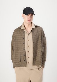 Brown and black checkered shirt jacket with a collar, front pockets, and black buttons over a beige button-up shirt, worn with a black cap.