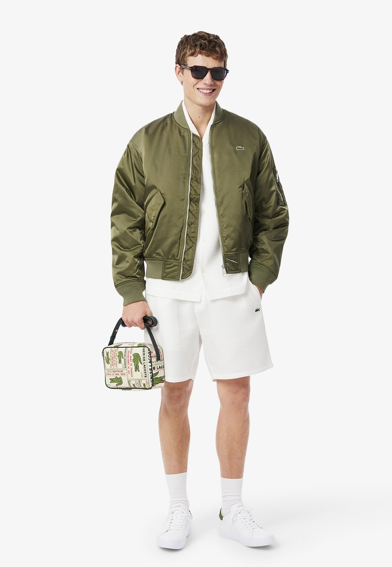 Giacca bomber in raso verde oliva, camicia bianca con colletto, pantaloncini bianchi strutturati, borsa stampata con manico nero. L'outfit è completato da sneakers bianche.