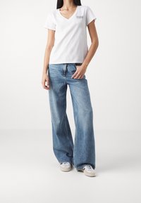 Femme portant un T-shirt Levi's blanc à col en V, un jean pattes d'éléphant bleu, et des baskets blanches, debout devant un fond clair uni.