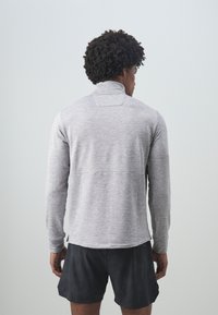 Grå långärmad sporttröja med ståkrage, tillverkad av texturerat tyg; ihop med svarta shorts som visar en avslappnad passform.