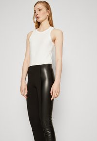 Bela rebrasta majica bez rukava u paru s crnim leggingsima visokog struka koji imaju gladak izgled kože. Model stoji na neutralnoj pozadini.