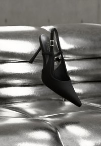 Scarpa slingback nera con punta a punta, finitura texturizzata, cinturino regolabile con fibbia e tacco stiletto, posizionata su una superficie riflettente.