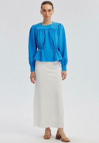 Blusa blu brillante con maniche a sbuffo e vita arricciata, abbinata a una gonna maxi bianca e texturizzata e sandali con tacco marroni.
