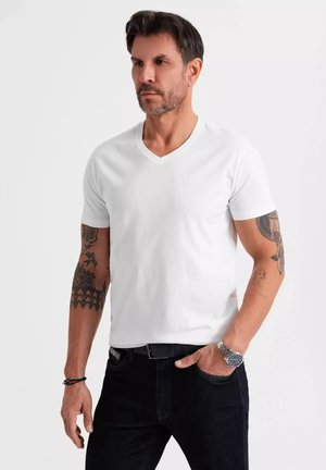 Camiseta blanca de manga corta y escote en V hecha de algodón suave. Presenta un ajuste relajado y se lleva con jeans de mezclilla azul oscuro y un reloj de pulsera plateado.