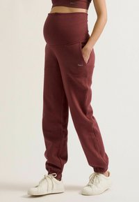 Zwangere joggers in bordeaux stof, met een hoge tailleband, zijzakken en geribbelde boorden, ontworpen voor comfort en bewegingsgemak.