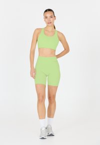 Une femme se tient face à l'avant, portant un soutien-gorge de sport vert citron et un short de cycliste taille haute assorti, avec des chaussettes blanches et des baskets grises.