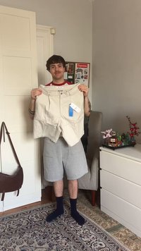 Shorts beiges retenus, avec une fermeture bouton et des plis, avec une étiquette bleue attachée. Shorts gris portés en dessous, se tenant sur un tapis à motifs.