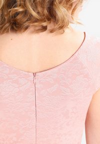 Lockigt hårig person som bär en ljusrosa klänning med blommönster, sedd från ryggen och axeln med en centrerad dragkedja.