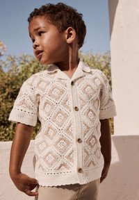 Nuori poika, joka pukeutunut valkoiseen virkattuun lyhythihainen nappipaitaan, seisoo ulkona vihreän kasvillisuuden ja rakennuksen edessä.