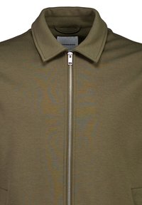 Olivgrüne Jacke mit strukturiertem Kragen, Reißverschluss vorne und strukturiertem Stoff. Besitzt ein klares Design und eine minimalistische Ästhetik.