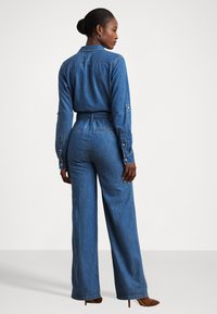 Combinaison en denim avec manches longues, taille ceinturée, coupe évasée et poignets boutonnés, dans une teinte bleue moyenne avec une texture lisse.