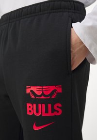 Sorte joggebukser med en tettsittende midje, med et rødt grafikk av en okse og teksten "BULLS" sammen med Nike-logoen i rødt.