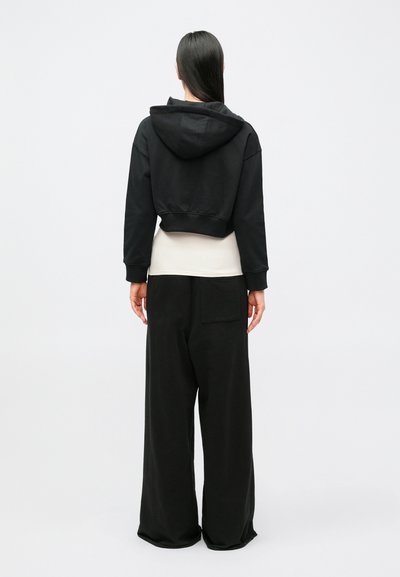 Sweatshirt preta com capuz, bainha canelada e mangas longas, usada sobre uma camisola clara; combinada com calças pretas de perna larga com um bolso traseiro.