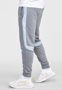 Graue Sweatpants mit hellblauen Seitenstreifen, die ein wiederholtes Logo-Muster aufweisen. Die Bündchen und Taschen sind in das Design integriert.