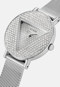 Reloj de pulsera de malla plateada con una caja triangular cubierta de brillantes cristales transparentes y una esfera minimalista, con el nombre de la marca "GUESS."