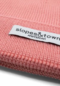 Różowa dziana czapka z prążkowanym wzorem, z białą metką z napisem "slopes&town AMSTERDAM" w czarnym foncie.