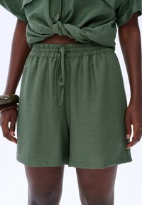 Vrouw in olijfgroene shorts met trekkoord en een korte mouw top die vastgebonden is, met armbanden om haar linkerpols.