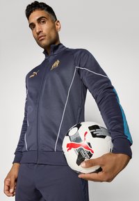 Chaqueta deportiva azul marino con detalles en blanco, cremallera en la parte delantera y logo de Puma. Una pelota de fútbol con un diseño está sostenida en la mano.