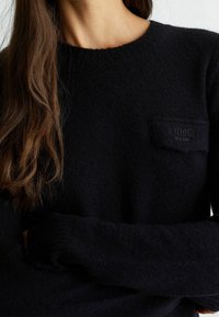 Svart stickad tröja med rund halsringning, mjuk textur och en bröstficka med en präglad logotyp. Långa ärmar med ribbade manschetter.