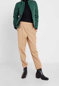 Giacca imbottita verde a sacco, maglione nero a costine, pantaloni beige plissettati e stivaletti neri. Design semplice, texture funzionale e colori tenui.