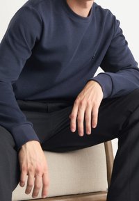 Námořnická modrá mikina vyrobená z měkké tkaniny, s kulatým výstřihem a žebrovanými manžetami, nošena přes černé kalhoty. Ruce odpočívají na světle zbarvené židli.