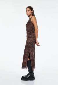 Frau in einem ärmellosen braunen Tiermusterkleid mit seitlichem Schlitz und schwarzen knielangen klobigen Stiefeln steht seitlich vor einem weißen Hintergrund.