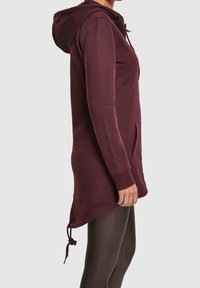 Femme portant un sweat à capuche long bordeaux avec poche kangourou et cordon de serrage, associé à des leggings foncés, vue de profil.
