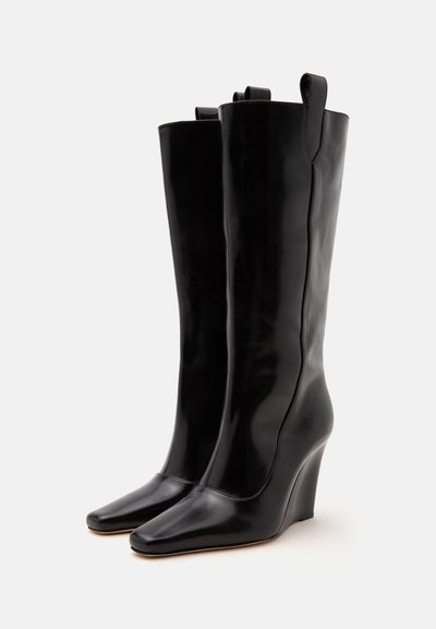 Paire de bottes compensées hautes en cuir noir avec bouts carrés et languettes de tirage en haut, exposées sur un fond clair uni.