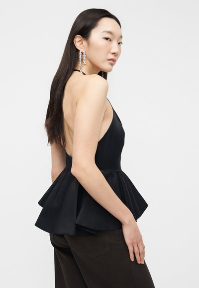 Femme aux longs cheveux foncés portant un haut noir sans manches à col licou avec un ourlet évasé à basque et des boucles d'oreilles en cristal pendantes, debout de profil.