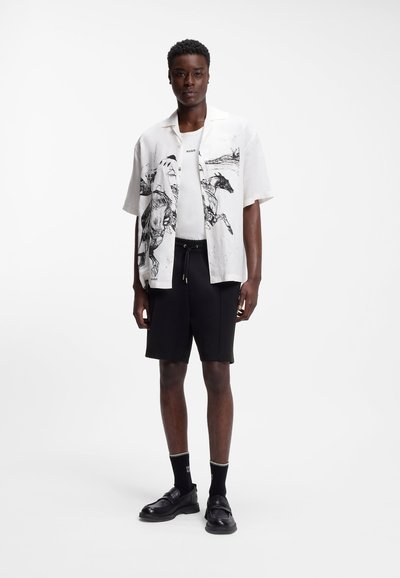 Homme portant une chemise à manches courtes blanche avec des imprimés de chevaux noirs, un T-shirt blanc, un short noir, des chaussettes noires et des mocassins noirs, debout devant un fond blanc.