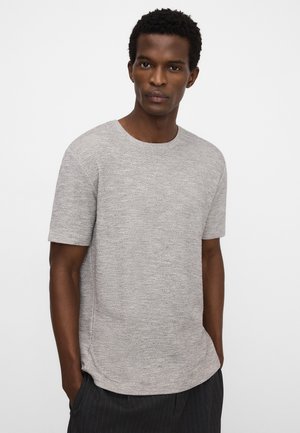 Homme aux cheveux courts et bouclés portant une chemise texturée à manches courtes gris clair et un pantalon sombre à fines rayures, mains dans les poches, fond neutre.
