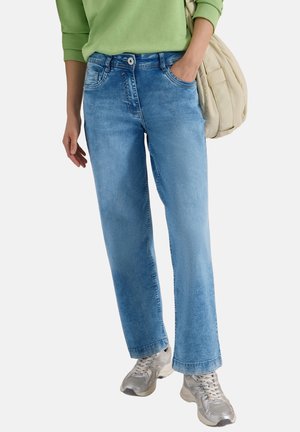 Personne portant un jean droit bleu clair, des baskets argentées, un haut vert à manches longues et un sac à épaule beige.