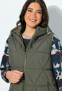 Usměvavá žena s tmavými vlasy, oblečená v zelené prošívané vestě přes vzorovanou košili s dlouhým rukávem, drží zip vesty u hladké zdi.