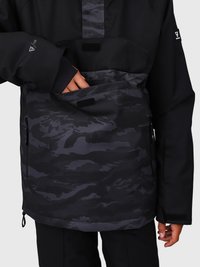 Persona vistiendo una chaqueta de camuflaje negra y gris con la mano dentro del bolsillo frontal con cremallera, de pie contra un fondo liso de color gris claro.