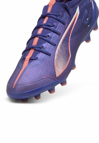 Fialové futbalové kopačky s textúrovaným vrchom, kontrastnými oranžovými akcentmi a aerodynamickým dizajnom. Obsahujú šnurovací uzáver a tvarované kolíky.
